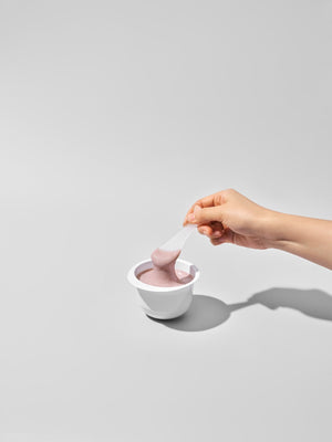 A hand holding a spatula scooping light purple skincare product from a white container, showcasing the creamy texture of 3X Boosting Modeling Mask - Collagen | Leaders.