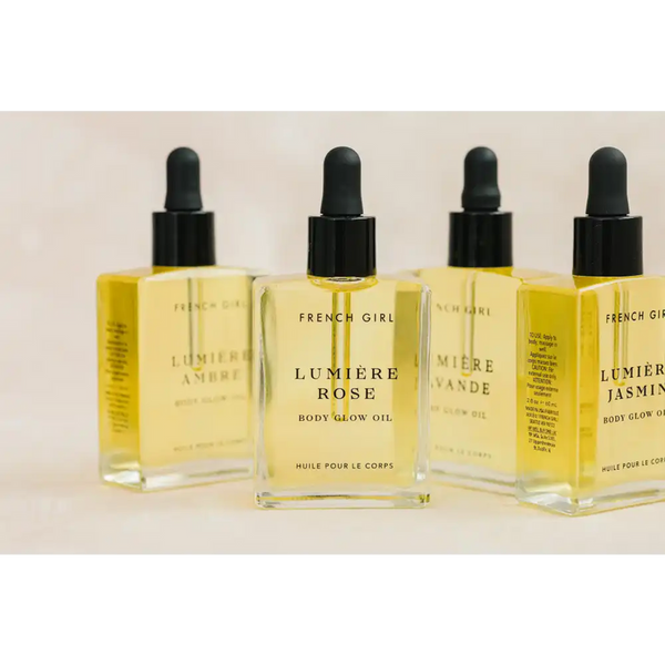Clear glass bottle of Lumière Body Glow Oil - Rose | French Girl with black dropper cap, surrounded by other natural body glow oil varieties, displayed on a neutral background.