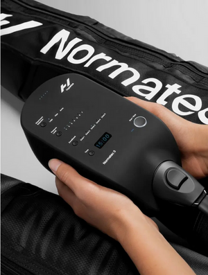 Hands holding a control unit of the Normatec 3 Leg Recovery System | Hyperice with a close-up view of the device and part of the black recovery leg sleeve visible in the background.