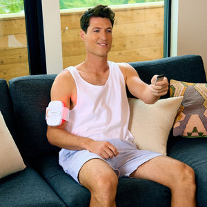 Man sitting on couch wearing a Recovery Loop | LightStim device on his upper arm while holding a remote control, demonstrating a skincare and wellness recovery tool at home.