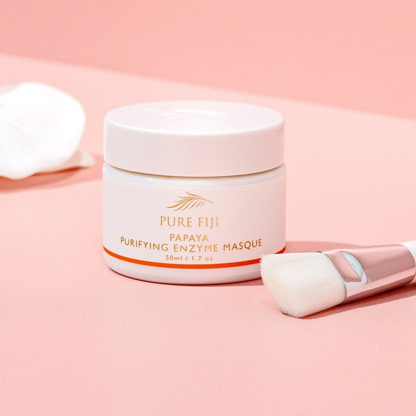 A jar of Papaya Purifying Enzyme Masque | Pure Fiji on a pink surface, accompanied by a soft cosmetic brush and a white cloth, highlighting skincare and wellness.