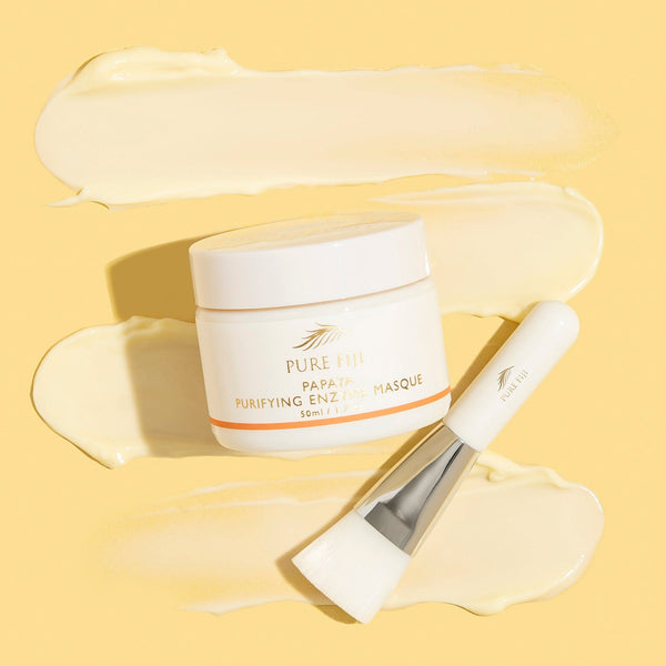 A jar of Papaya Purifying Enzyme Masque | Pure Fiji with a white applicator brush resting on a smooth, creamy pale yellow skincare masque texture background.