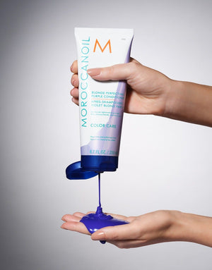Hand holding a tube squeezing vibrant purple conditioner onto the other palm with the product name Blonde Perfecting Purple Conditioner | Moroccanoil visible on the tube.