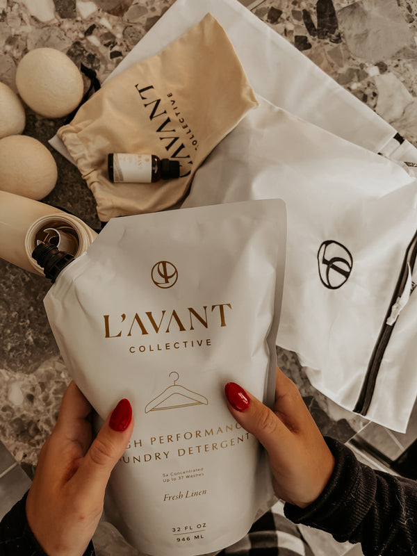 Hands with red nail polish hold a white Laundry Detergent Refill Pouch | L'AVANT Collective above a marble surface with beige and white cloth bags and natural laundry accessories.