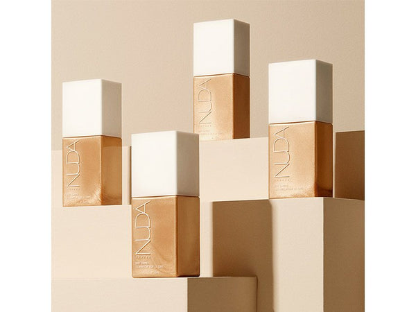 Four gold-toned Body Shimmer | NUDA bottles with white caps displayed on beige geometric platforms, highlighting skincare and body care luxury.