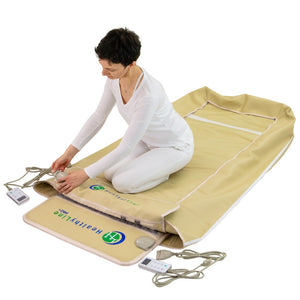 A woman in white clothing kneeling next to a beige wellness device labeled TAJ-Mat™ Full 7224 Firm - Photon PEMF InfraMat Pro® | HealthyLine, with control units and cords visible nearby.