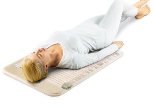 Woman lying relaxed on a beige wellness mat designed for body care and recovery, showcasing the SOFT Mat Medium 4024 InfraMat Pro® | HealthyLine in use.