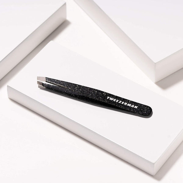 Anniversary Slant Tweezer | Tweezerman in black with a speckled design, displayed on a white geometric platform, ideal for precise eyebrow grooming in skincare routines.