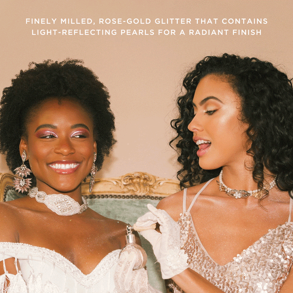 Two women wearing elegant dresses and jewelry smile while one applies Glitter Dust | Pretty Vulgar rose-gold glitter makeup with light-reflecting pearls for a radiant finish