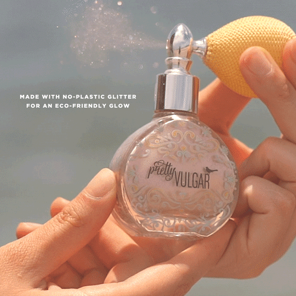 Person spraying sparkling eco-friendly glitter mist from a vintage-style glass bottle labeled Glitter Dust | Pretty Vulgar against a soft blue background.