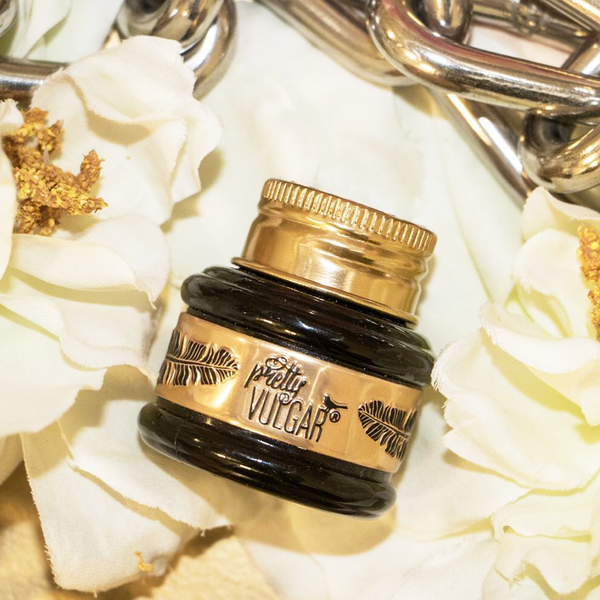 Close-up of a small black and gold jar of The Ink Gel Eyeliner | Pretty Vulgar surrounded by white flowers and gold chain links on a soft surface.