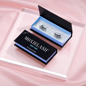 Black magnetic false eyelashes displayed in an open and closed box on a clear acrylic platform with a soft pink fabric backdrop, featuring the product name Baby Lash | Moxielash.