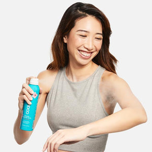 A smiling woman in a gray sleeveless top applying Classic Body Organic Sunscreen Spray SPF 50 - 6 fl oz | COOLA to her arm, demonstrating body care and skincare wellness.