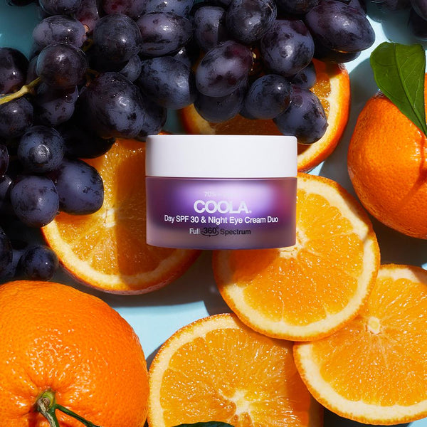 Jar of Full Spectrum 360° Day SPF 30 & Night Organic Eye Cream Duo | COOLA surrounded by fresh oranges and purple grapes, highlighting natural skincare ingredients.