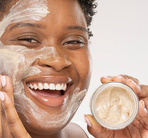 Smiling person with a creamy exfoliating scrub applied on their face holding an open jar of Finely Awake Plantfoliant Silkening Face Cleansing Polish | Farmhouse Fresh, highlighting skincare and self-care.