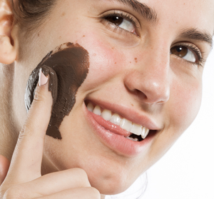 Close-up of a woman smiling while applying a dark brown facial mask on her cheek using her finger, showcasing skincare with the Fast Fix Face Mask Sampler | Farmhouse Fresh.
