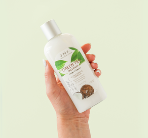 Hand holding a white bottle of Green Tea Milk Wash | Farmhouse Fresh with green tea leaves and natural ingredients illustration on the label against a light green background, highlighting skincare and body care.