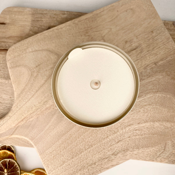 Top view of an unlit Santal + Coconut 10 oz Candle | ROAM Homegrown with a wooden wick, placed on a light wooden cutting board next to dried citrus slices.