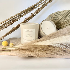 Two Santal + Coconut 10 oz Candle | ROAM Homegrown with natural beige containers, presented alongside dried grasses and decorative botanicals on a light surface.