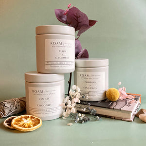 Three white ceramic jars of scented soy candles labeled Plum Cashmere, Pumpkin Cinnamon, and Santal Coconut arranged with dried flowers, a dried orange slice, and a sage bundle, promoting Lavender + Driftwood 10 oz Soy Candle | ROAM Homegrown.