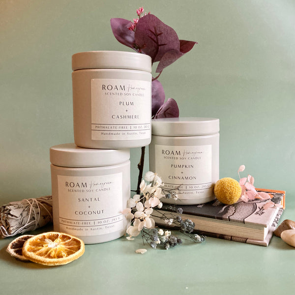 Three white ceramic jars of scented soy candles labeled Plum Cashmere, Pumpkin Cinnamon, and Santal Coconut arranged with dried flowers, a dried orange slice, and a sage bundle, promoting Lavender + Driftwood 10 oz Soy Candle | ROAM Homegrown.