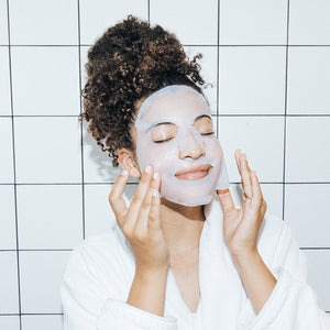 Woman wearing a white facial sheet mask and a white robe, gently touching her face with eyes closed, demonstrating skincare relaxation with LightStim SheetMasque | LightStim.