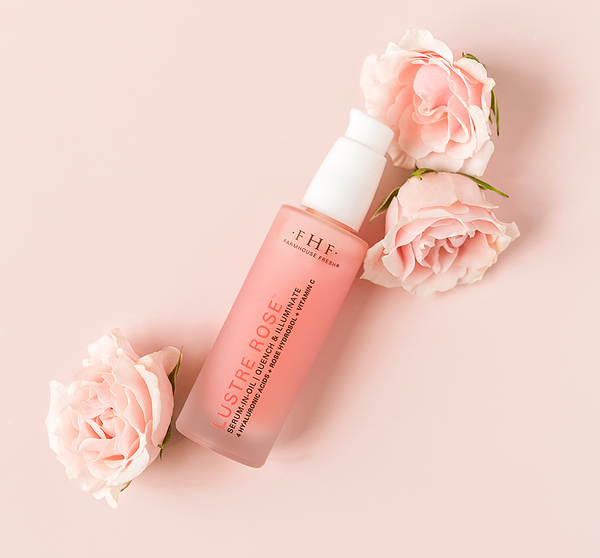 Lustre Rose™ Serum-in-Oil | Farmhouse Fresh skincare product bottle surrounded by soft pink roses on a pale pink background for a fresh and soothing wellness aesthetic.