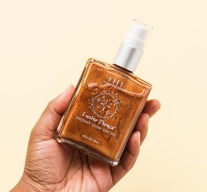 Hand holding a square glass bottle of Lustre Drench® Instant Glow Dry Oil | Farmhouse Fresh with shimmering bronze oil inside against a light beige background, showcasing skincare and body care product.