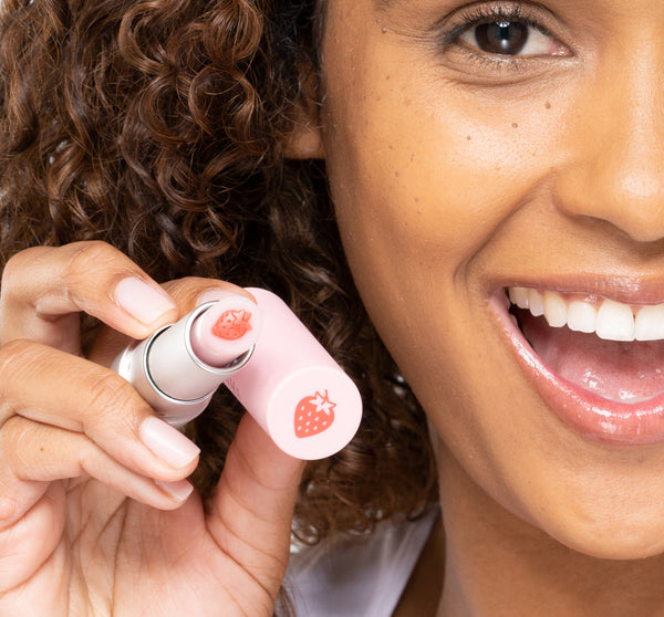 Smiling woman holding Strawberry Mood Fruit™ Lip Therapy | Farmhouse Fresh with visible strawberry design on the pink cap, showcasing skincare lip treatment.