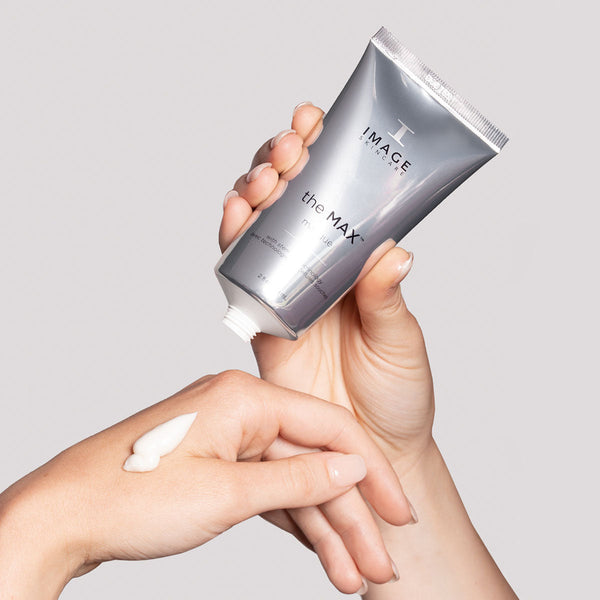 Hand applying creamy white skincare product from a silver tube labeled the MAX™ masque | IMAGE Skincare, demonstrating smooth texture and hydration.