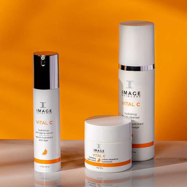 Three IMAGE Skincare products on a reflective surface with a vibrant orange background, featuring VITAL C hydrating repair crème | IMAGE Skincare alongside a serum and facial cleanser.